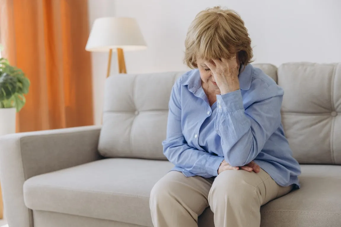 Eine ältere Frau sitzt auf dem Sofa und hält sich mit der Hand ihren Kopf