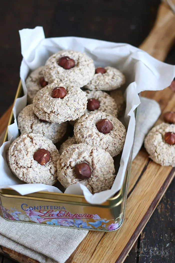 Eine Auswahl an fertig gebackenen Haselnussmakronen in einer Dose, einige Makronen sind gestapelt, während andere einzeln liegen. Die Makronen haben eine goldbraune Farbe und sind mit einer ganzen Haselnuss belegt.
