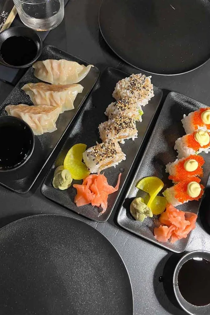 Eine Auswahl an Sushi und Gyoza, serviert auf schwarzen Keramiktellern, begleitet von Sojasauce, eingelegtem Ingwer, Wasabi und Zitronenscheiben, präsentiert auf einem dunklen Restauranttisch in einem der besten japanischen Restaurants in Zagreb.