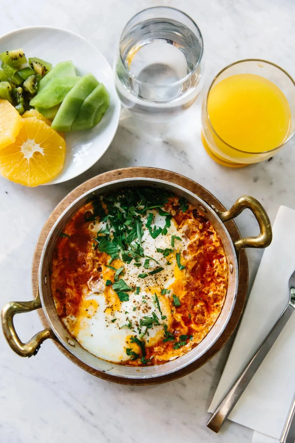 Eine Einzelportion Shakshuka in einer kleinen Pfanne, serviert mit frischem Orangensaft und einer Obstschale.