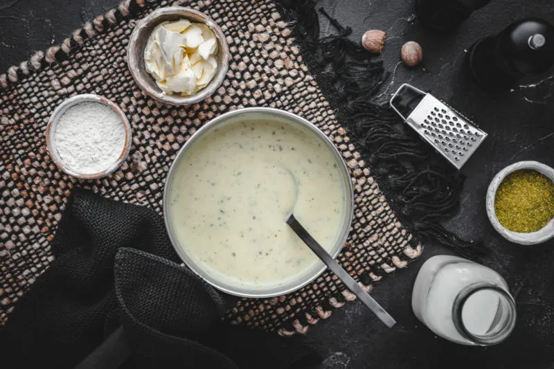 Eine elegante helle Schüssel, gefüllt mit perfekt zubereiteter Béchamelsoße. Um die Schüssel herum sind alle notwendigen Zutaten für die Soße, sorgfältig in kleinen Gefäßen angeordnet.