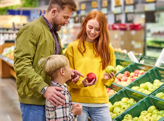 Eine Familie steht an einem Obstregal und vergleicht Äpfel.