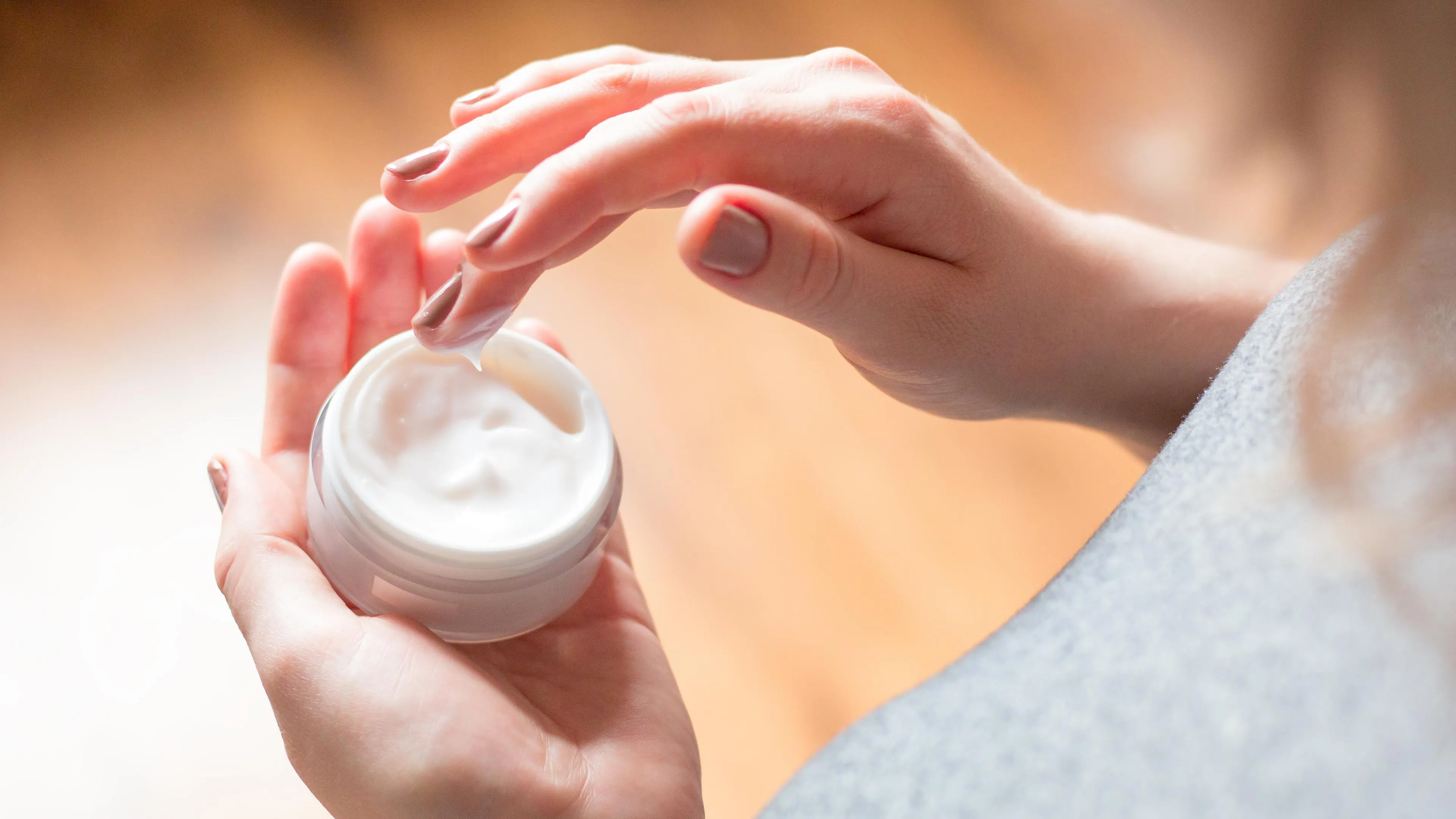 Eine Frau entnimmt vorsichtig Gesichtscreme aus einem Tiegel, Symbol für Hautpflege und Behandlungen