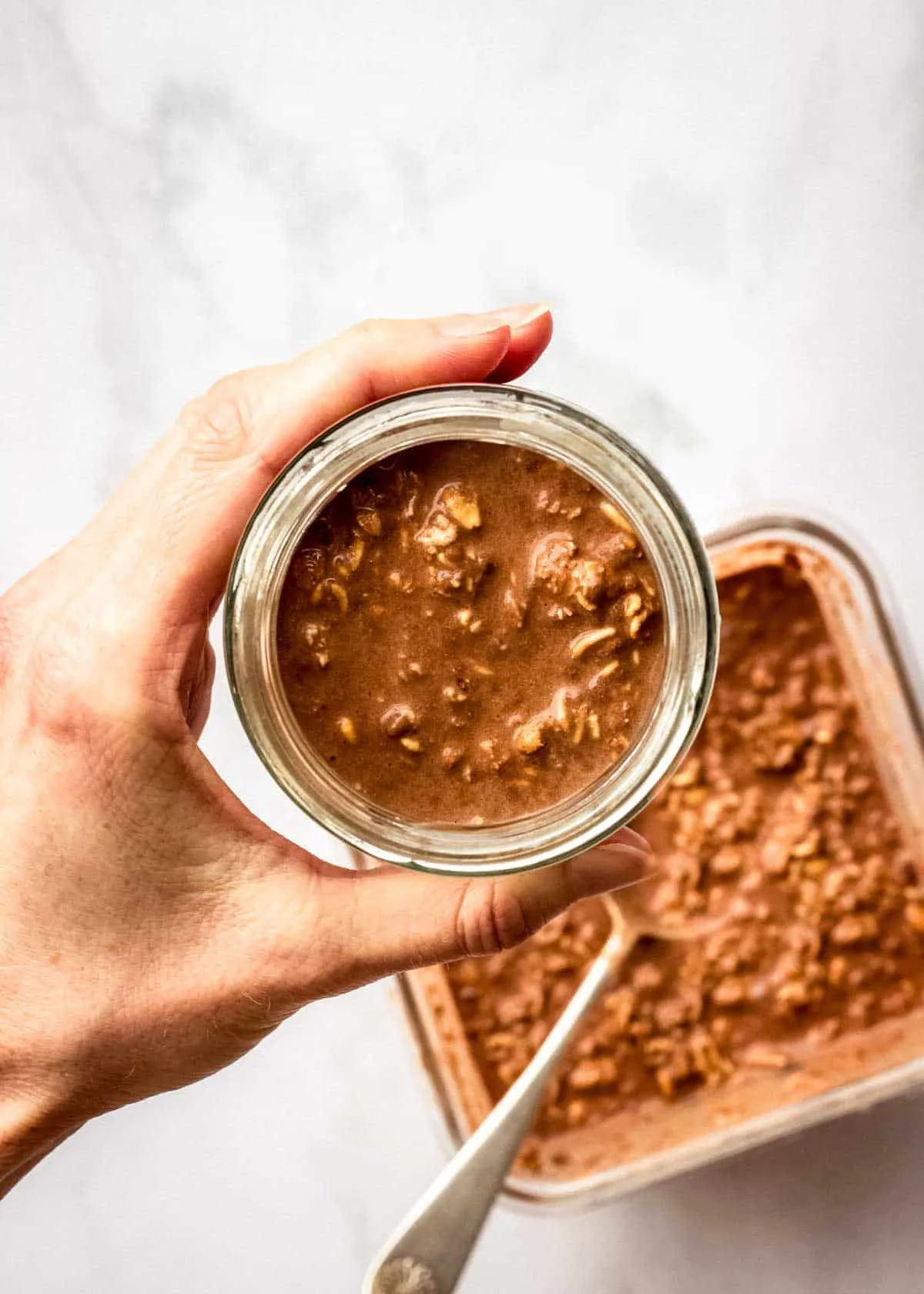 Eine Frau hält ein Glasgefäß mit Schokoladen-Espresso Overnight Oats. Ein Behälter mit der Haferflockenmischung ist im Hintergrund sichtbar.