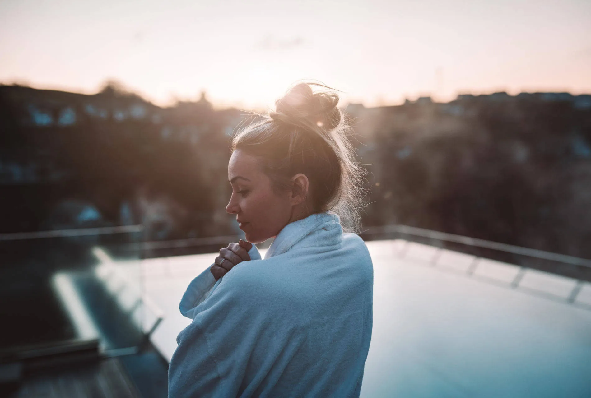Eine Frau steht auf der Terrasse neben dem Außenpools und kuschelt sich in ihren Bademantel