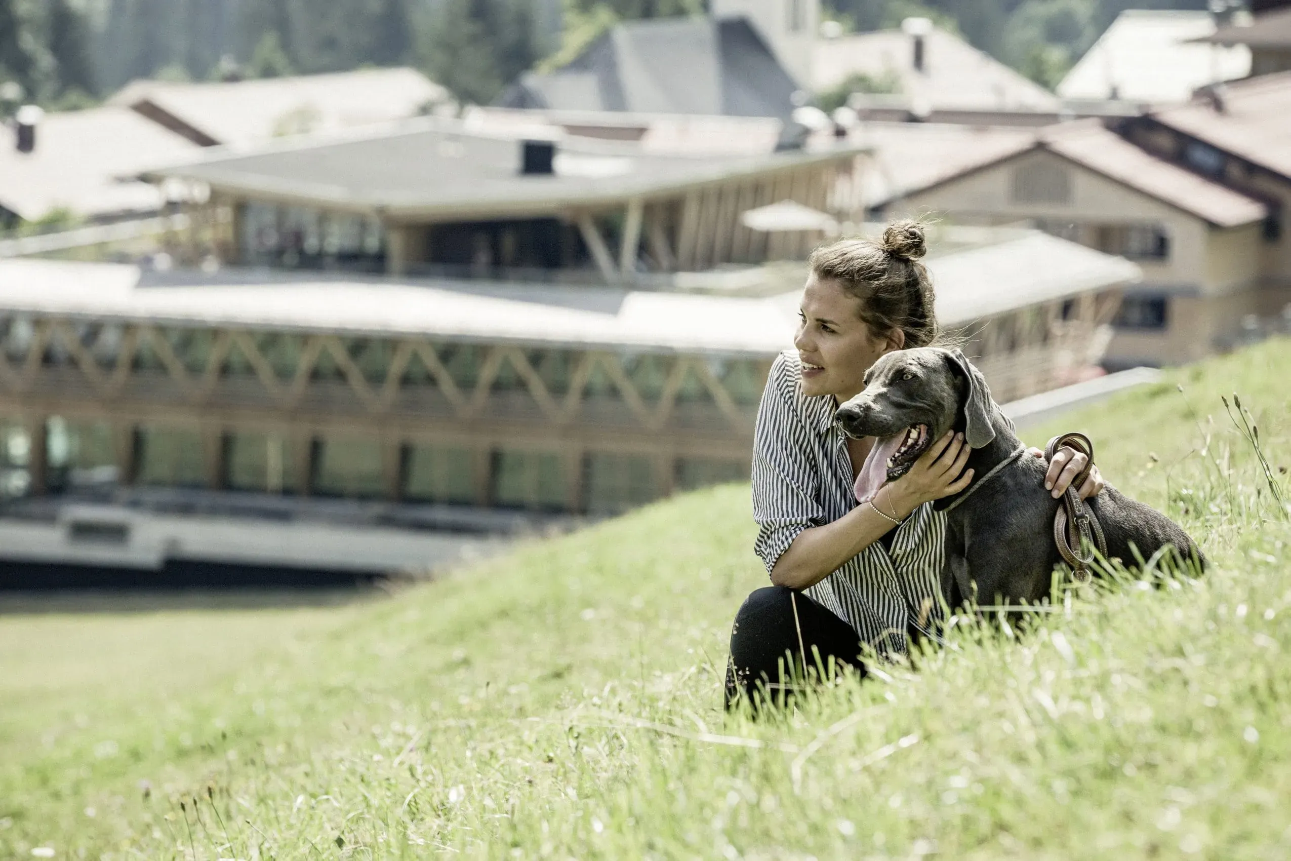 Eine glückliche Frau spielt mit ihrem Hund auf einer grünen Wiese vor dem malerischen HUBERTUS Mountain Refugio Allgäu, einem idealen Hotel mit Hund für einen erholsamen Urlaub.