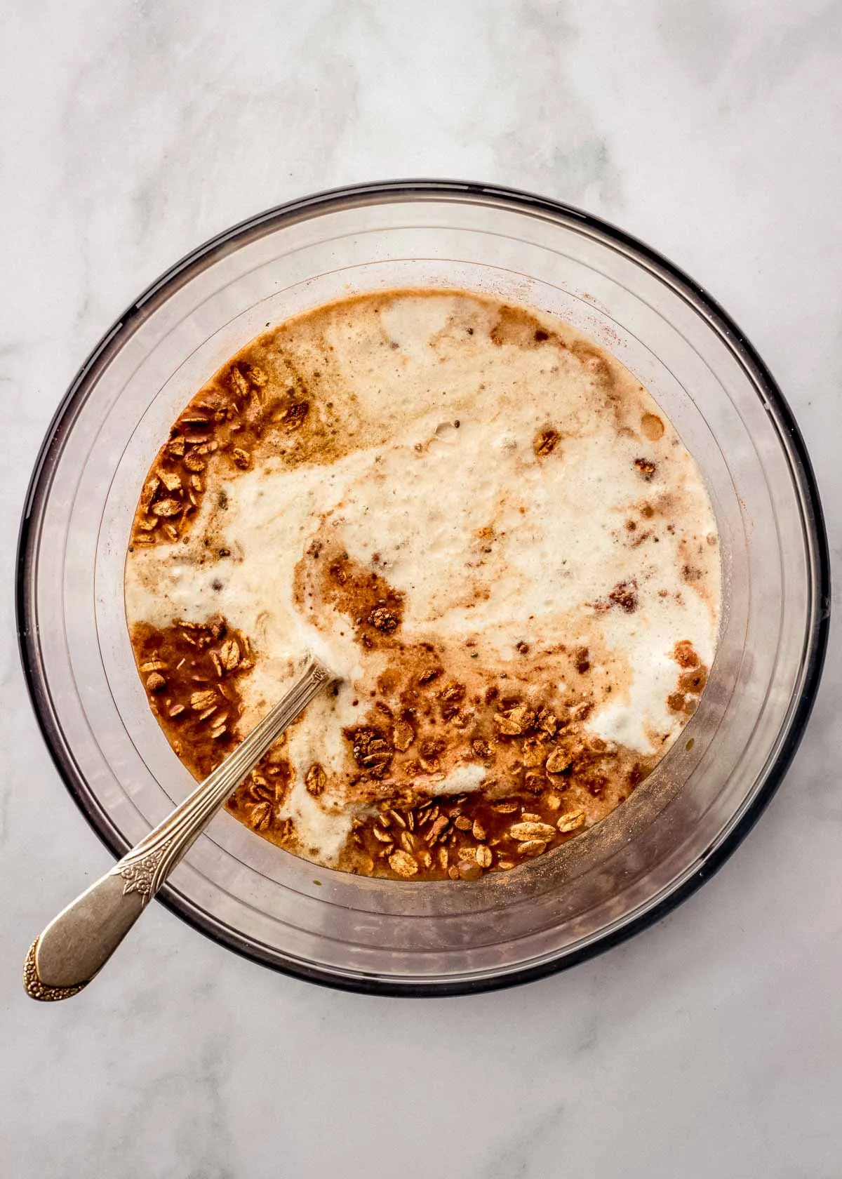 Eine große Glasschüssel mit Tiramisu Overnight Oats Zutaten, ein Löffel darin, bereit zum Umrühren.