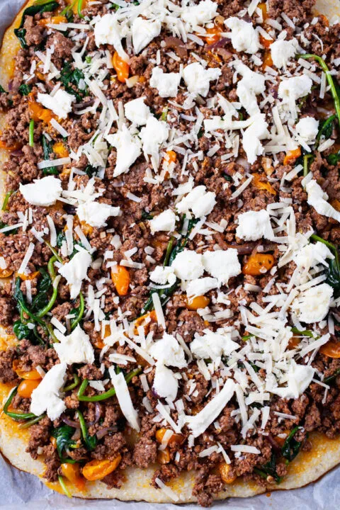 Eine große glutenfreie Polenta Pizza, belegt mit gebratenem Hackfleisch, Gemüse und Mozzarella-Käse, bereit zum Backen im Ofen.