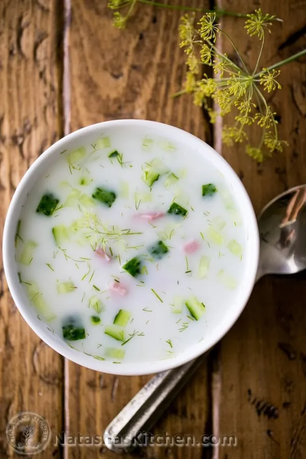 Eine große Schale mit Okroschka auf einem Holztisch, umgeben von frischen Zutaten und bereit für eine Mahlzeit.