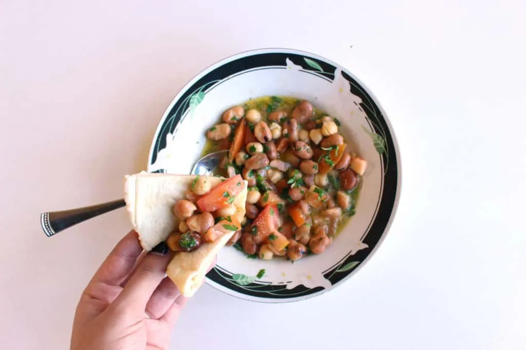 Eine Hand hält einen Löffel mit cremigem Ful Medames, dem syrischen Bohnensalat, frisch zubereitet und servierbereit, als Teil eines gesunden Frühstücks.