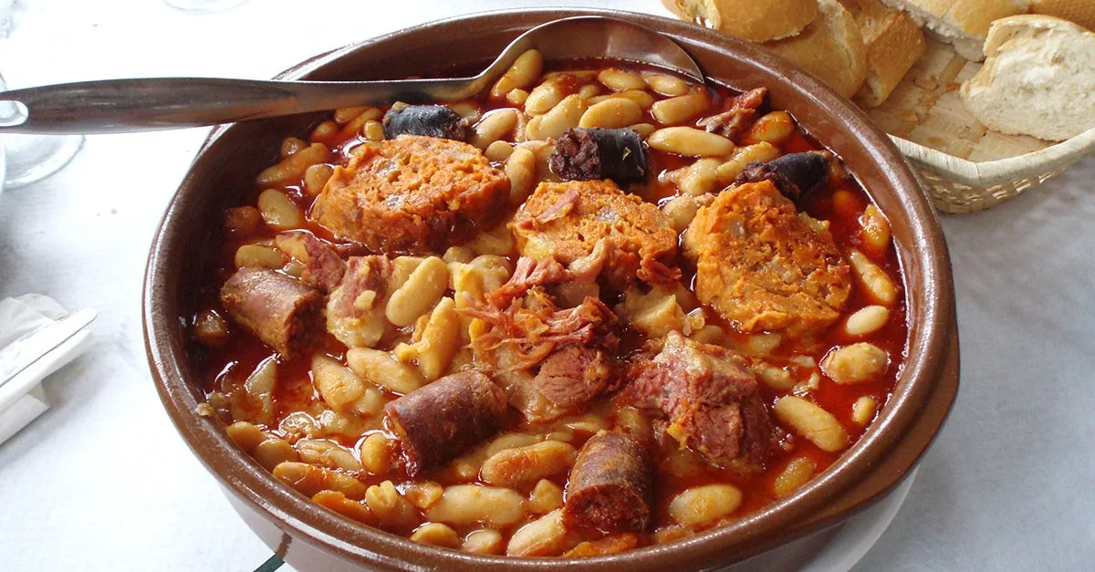 Eine herzhafte asturische Fabada, ein reichhaltiger weißer Bohneneintopf mit Chorizo, Morcilla und Schweinefleisch, dampfend in einer rustikalen Keramikschale