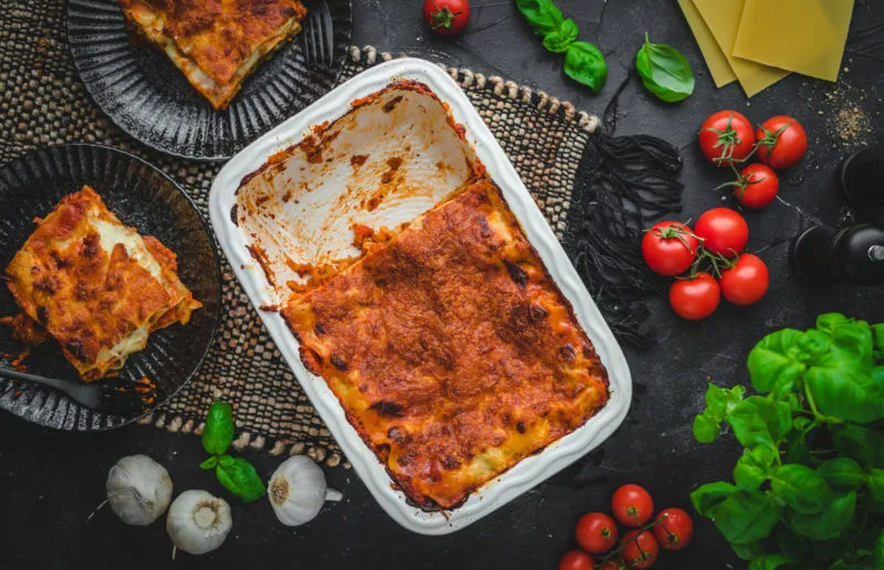 Eine klassische weiße Auflaufform, randvoll gefüllt mit einer appetitlichen vegetarischen Lasagne, platziert auf einem dunklen Hintergrund. Daneben befinden sich dekorative Elemente und zwei schwarze Teller, die jeweils ein Stück Lasagne präsentieren.