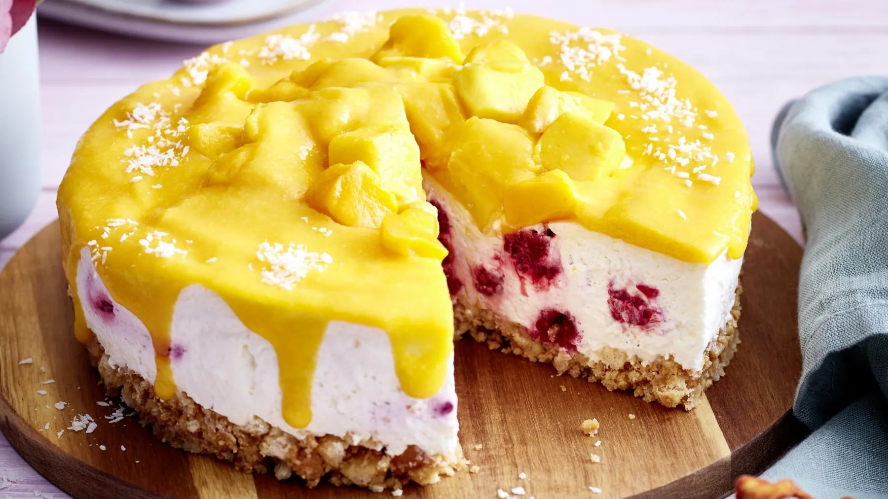 Eine Mango-Himbeer-Torte auf einem hölzernen Tortenteller. Rechts davon ein graues Geschirrtuch.