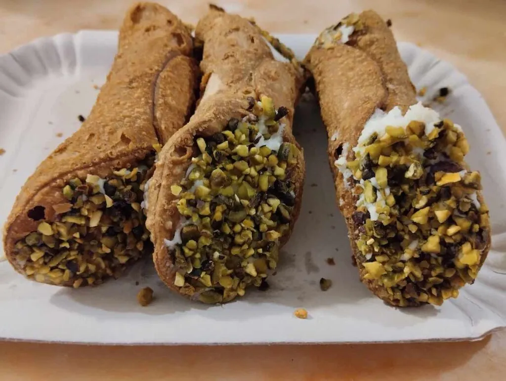 Eine Platte mit Cannoli, deren knusprige Hüllen in Schokolade und Pistazien getaucht und mit cremiger Ricotta gefüllt sind, wartet auf einem weißen Pappteller und lockt mit sizilianischer Süße.