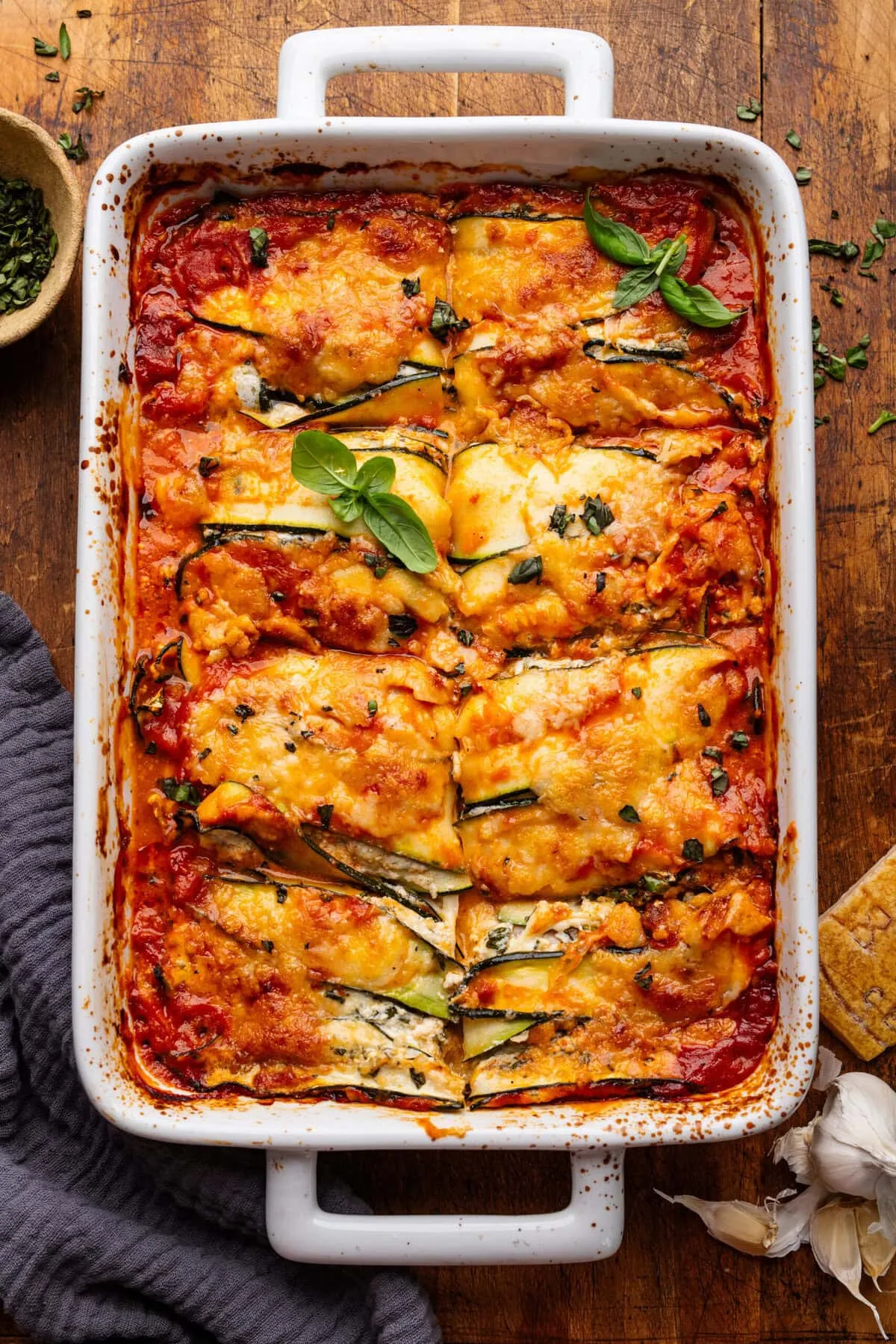Eine rechteckige weiße Auflaufform mit Zucchini Lasagne auf einem Holzbrett, umgeben von Kräutern, einer dunklen grauen Serviette, Knoblauchzehen und einer Käsrinde.