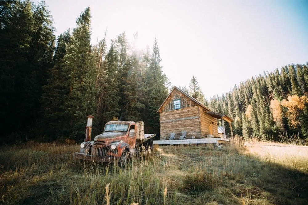 Eine rustikale Holzhütte und ein alter, rostiger Pickup-Truck stehen auf einer grasbewachsenen Lichtung in Colorado, umgeben von hohen Kiefern im hellen Sonnenlicht.