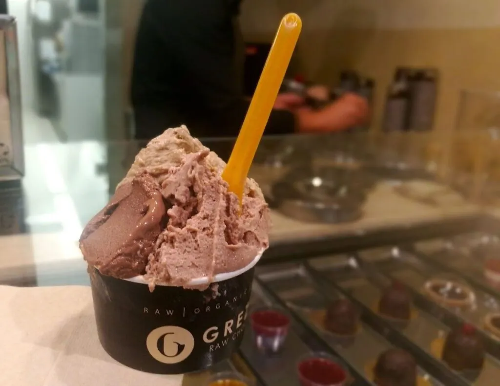 Eine Tasse veganes Gelato von der römischen Gelateria Grezzo mit einer Kugel Schokolade und einer Kugel Haselnuss, serviert mit einem leuchtend gelben Löffel, eine einladende Leckerei für einen milchfreien Genuss.