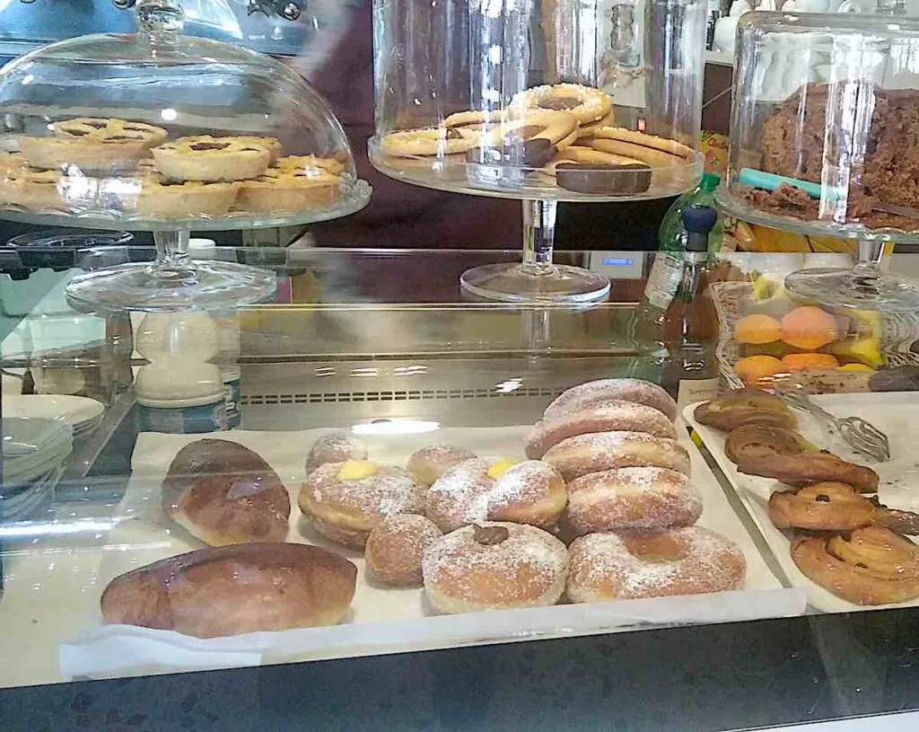 Eine Vitrine in einer römischen Bäckerei mit einer Auswahl an frisch gebackenem Gebäck, darunter mit Zucker bestäubte Bomboloni, Schokoladentörtchen unter einer Glaskuppel und flockige, mit Schokolade gefüllte Gebäckstücke.