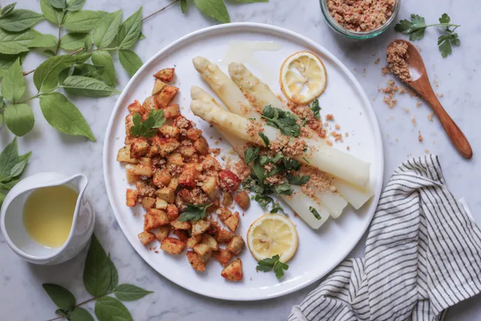Fertig angerichteter weißer Spargel mit veganen Butterbröseln, Ofenkartoffeln und frischer Petersilie, ein Festmahl.