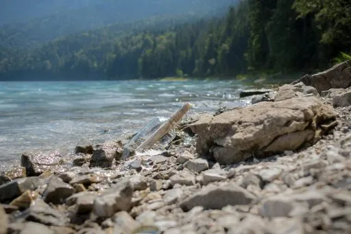 Flaschenpost mit Papier im klaren Wasser eines Bergsees, umgeben von Felsen und Wald im Hintergrund