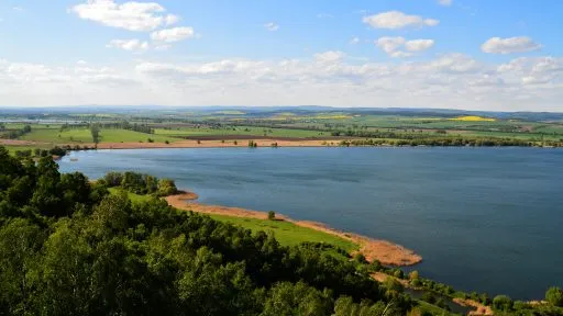 Fluss mit breitem Wasserlauf, umgeben von grünen Wiesen und Feldern unter blauem Himmel mit Wolken