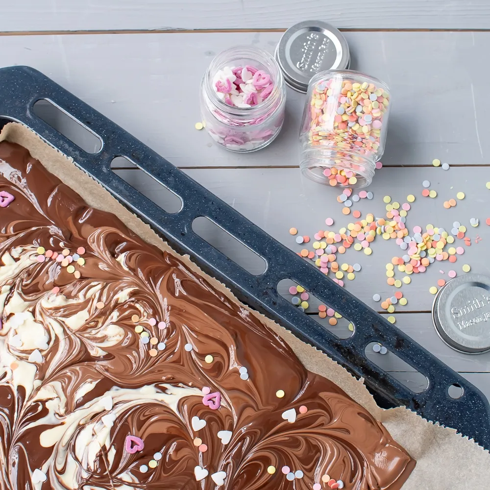 Frisch geschmolzene und verzierte Bruchschokolade mit Zuckerherzen und Konfetti auf dem Backblech, bereit zum Abkühlen und Brechen.