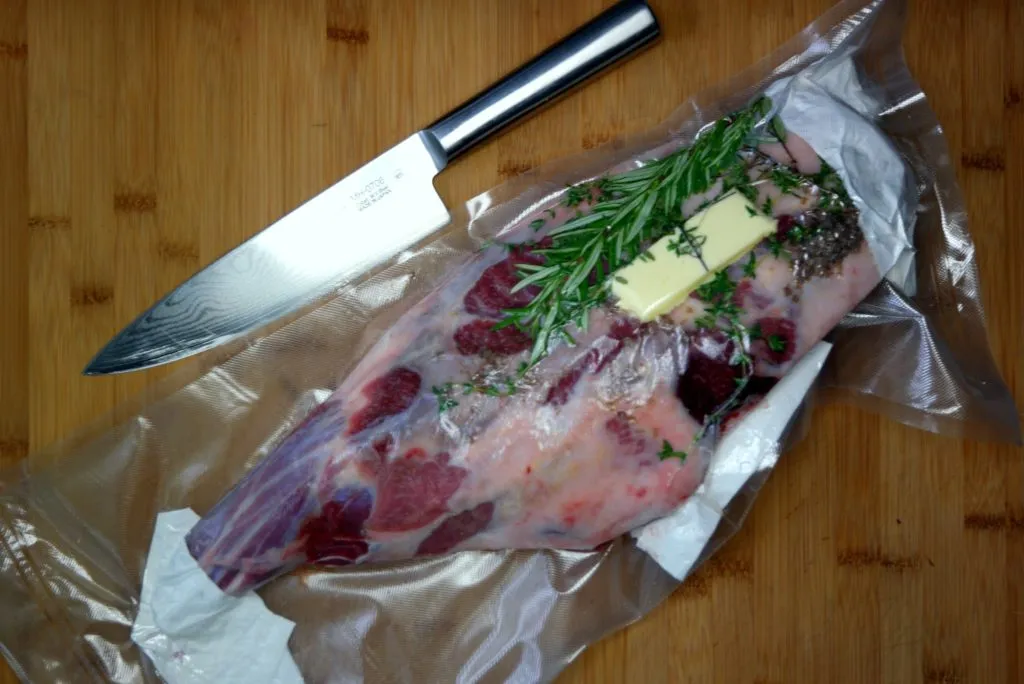 Frische Kräuter wie Rosmarin und Thymian und Butter auf der vorbereiteten Lammkeule vor dem Vakuumieren
