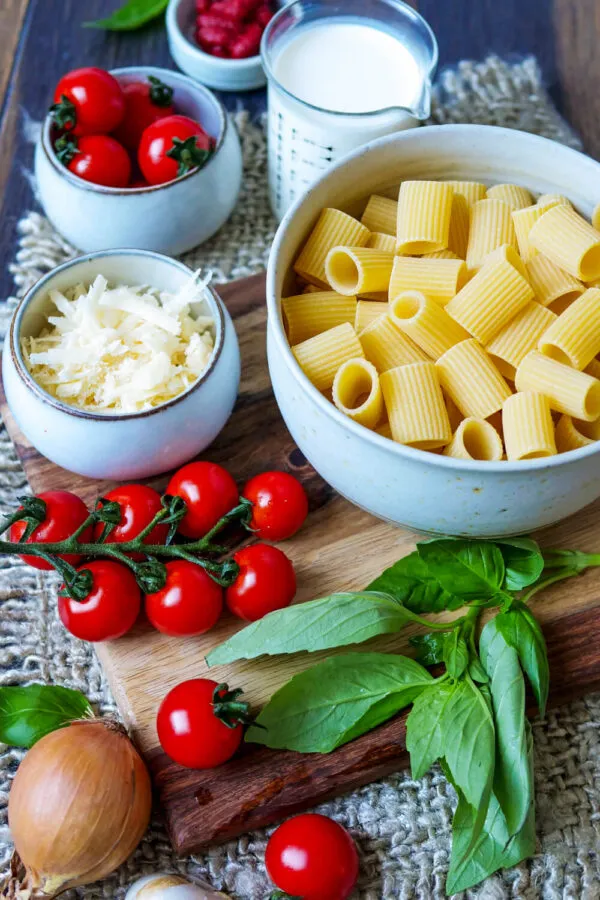 Frische Zutaten für die schnelle One Pot Pasta auf einem Holzbrett: Nudeln, Kirschtomaten, Basilikum, Zwiebel und Knoblauch bereit zur Vorbereitung.