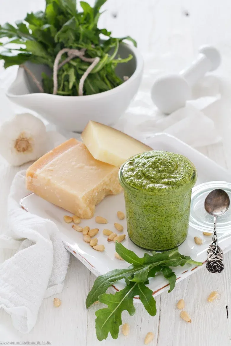 Frisches Rucola Pesto auf einem Holztisch, umgeben von seinen Zutaten wie Parmesan, Pinienkernen und Olivenöl.