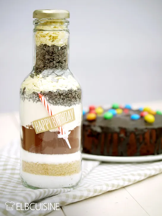 Geburtstagskuchen Backmischung im Glas mit Schichten von trockenen Zutaten, dekoriert mit Band und Etikett für ein einzigartiges Geschenk.