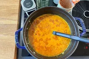 Gemüsebrühe und gehackte Tomaten werden zu den Linsen und Gewürzen im Topf gegeben und zum Köcheln gebracht, Basis für eine würzige Linsensuppe.