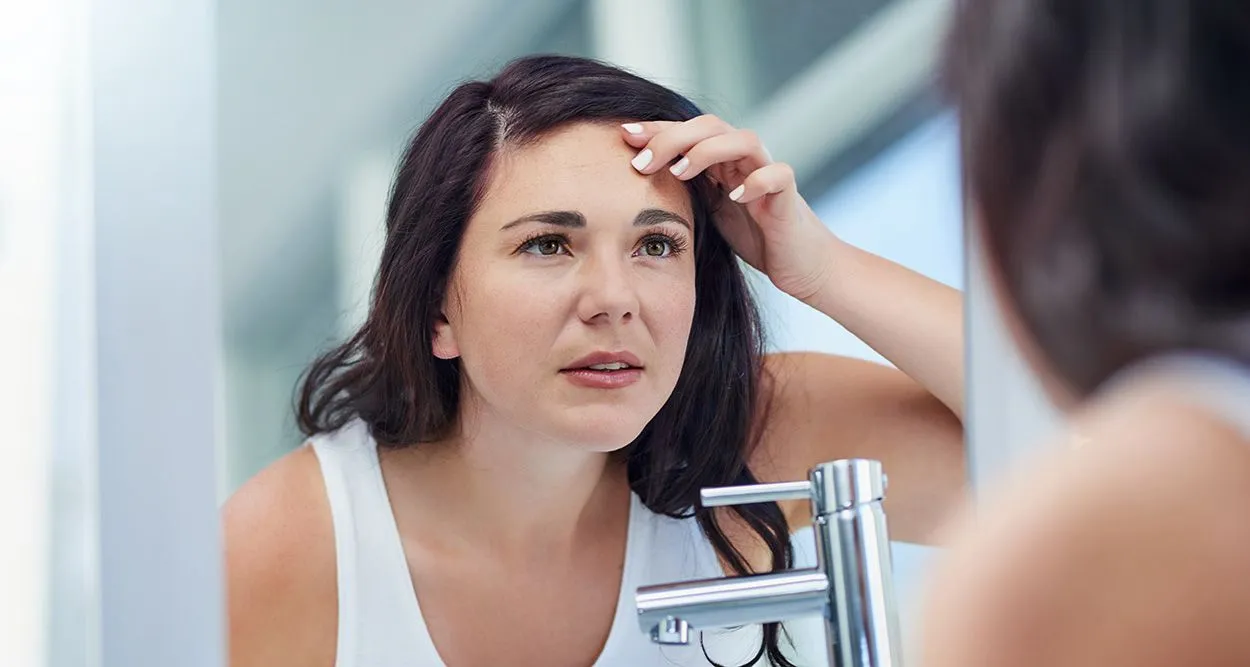 Gesicht einer jungen Frau mit einigen Pickeln und Rötungen, die sich besorgt im Spiegel betrachtet