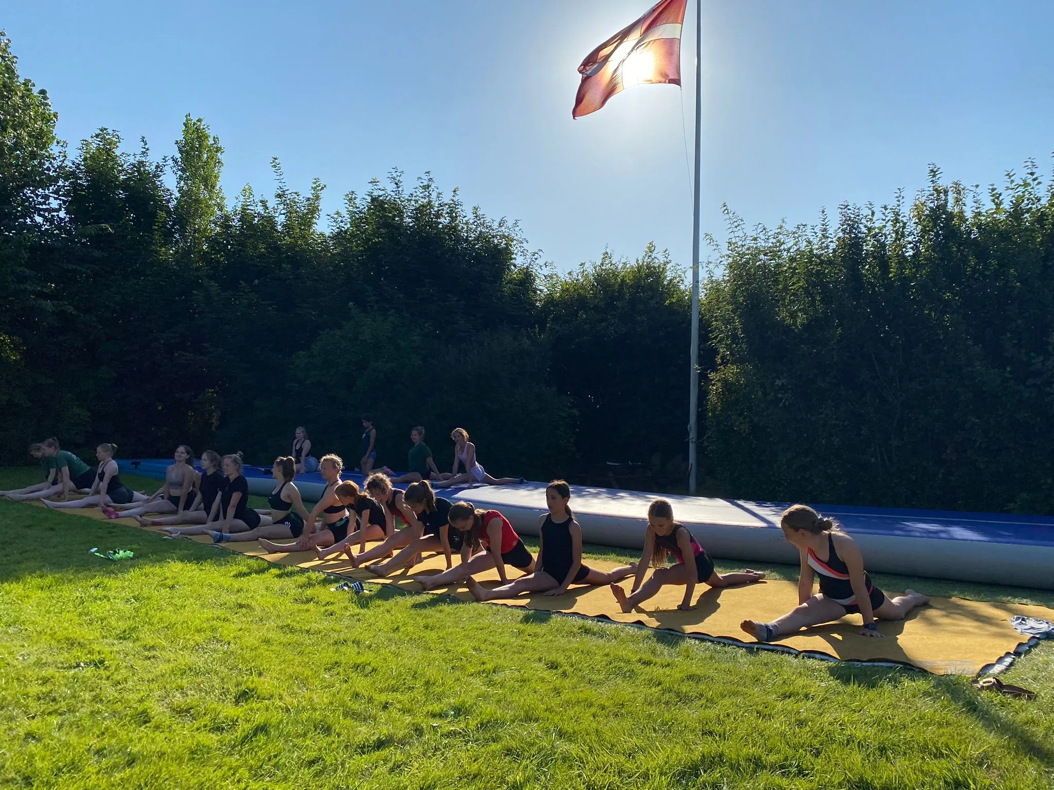 Gruppe von Turnerinnen trainiert an verschiedenen Geräten im Freien, darunter eine Schnitzelgrube und ein Trampolin