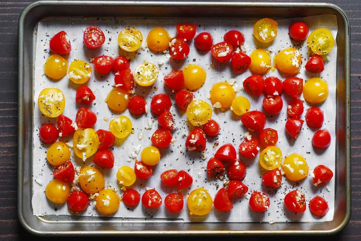 Halbierte Kirschtomaten und gehackter Knoblauch mit Olivenöl vermischt auf einem Backblech, bereit zum Rösten.