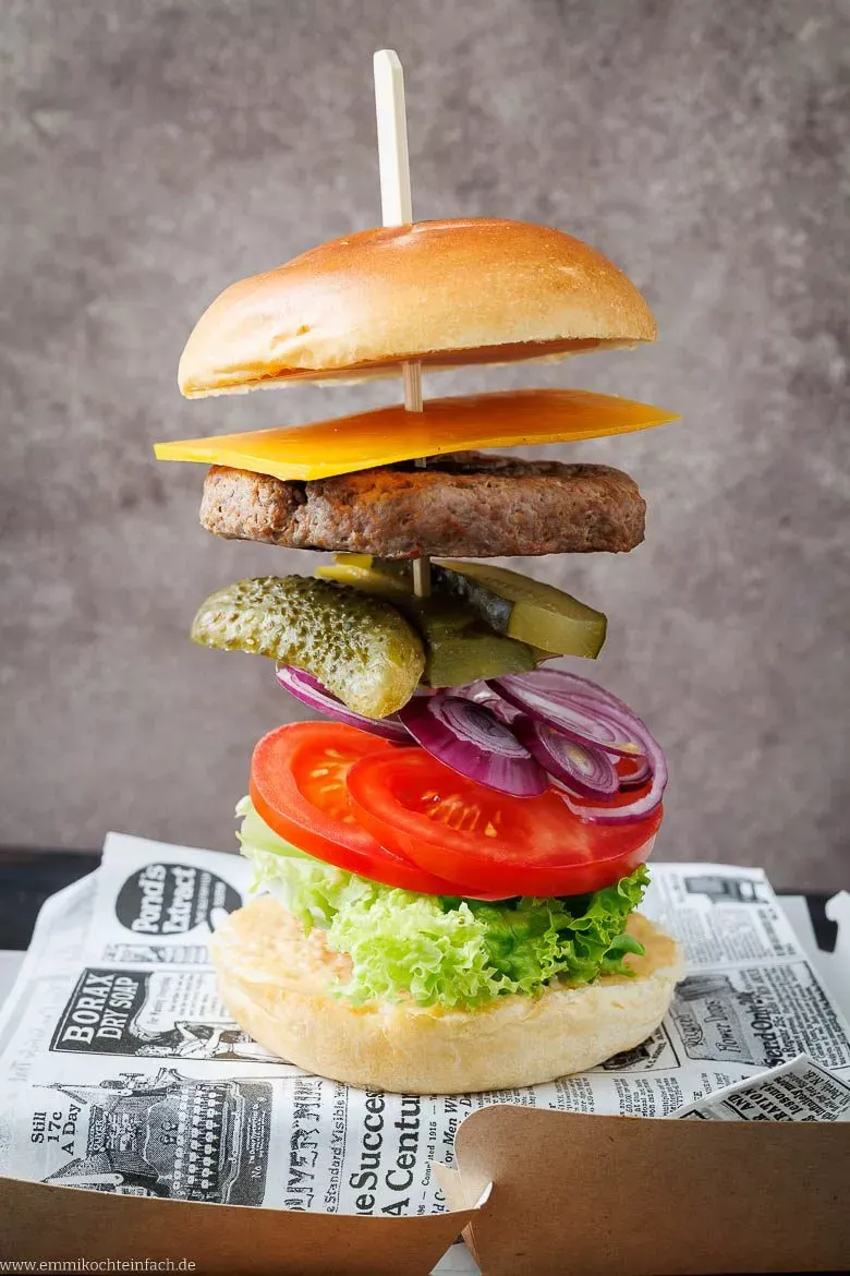 Hände belegen einen Burger Schritt für Schritt: Von der Sauce auf dem Brötchen bis zum saftigen Patty, Käse und frischen Gemüsesorten.