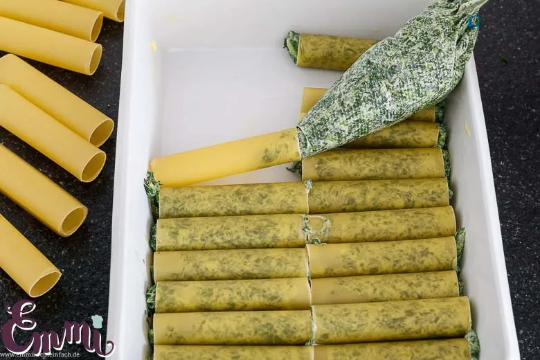 Hände füllen ungekochte Cannelloni-Röllchen mit der Spinat-Masse direkt in eine Auflaufform.