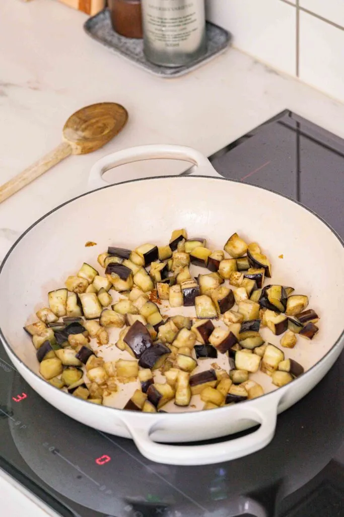 In Würfel geschnittene Auberginen, die in einer weißen Pfanne auf einem Elektroherd gebraten werden, mit einem Holzlöffel auf der Theke in der Nähe.
