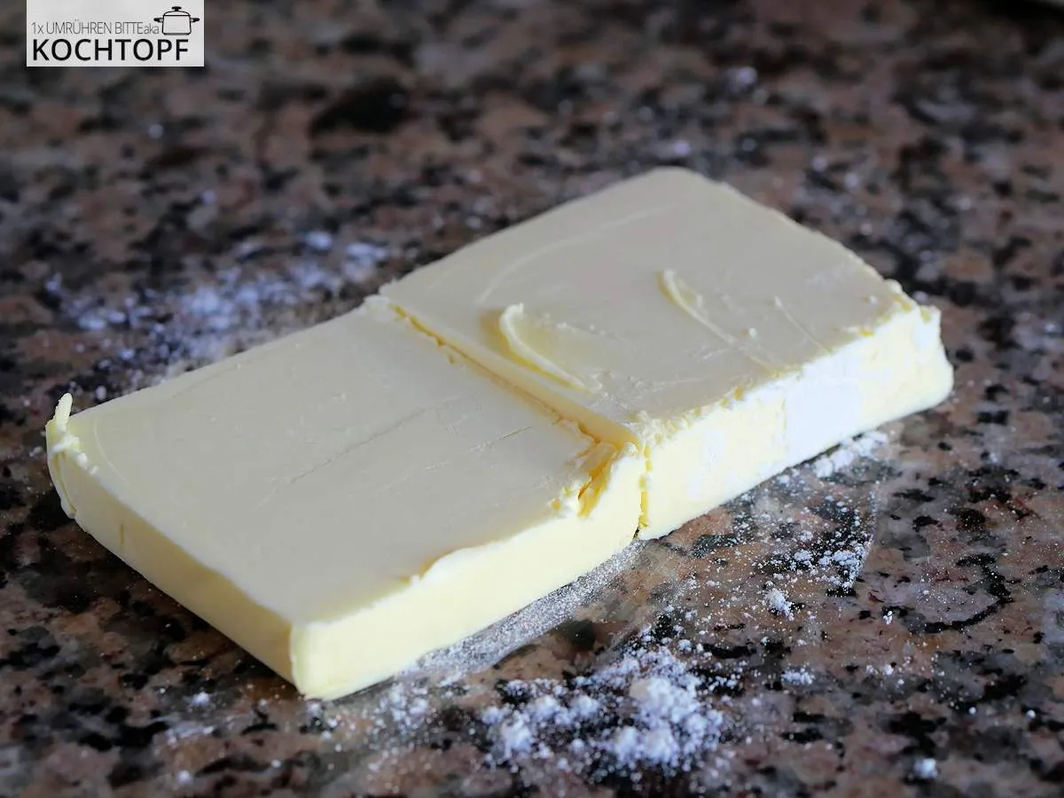 Kaltes Butterstück, längs halbiert auf einer leicht bemehlten Arbeitsfläche, vorbereitet für die Blätterteig-Zubereitung.