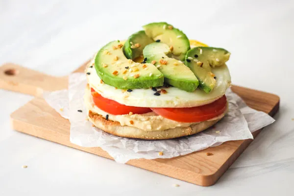 Kleines Schneidebrett mit einem English Muffin, belegt mit Hummus, Ei und Avocadoscheiben, perfekt für ein schnelles Toaster-Frühstück.