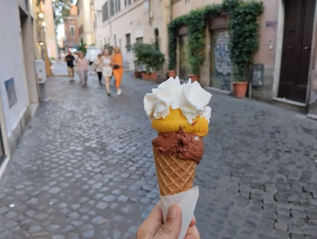 Kugeln aus reichhaltiger Schokolade und lebhaftem Mango-Gelato, garniert mit Schlagsahne in einer knusprigen Waffel, vor der Kulisse einer Kopfsteinpflasterstraße in Rom, die das Wesen italienischer Sommerleckereien einfängt.