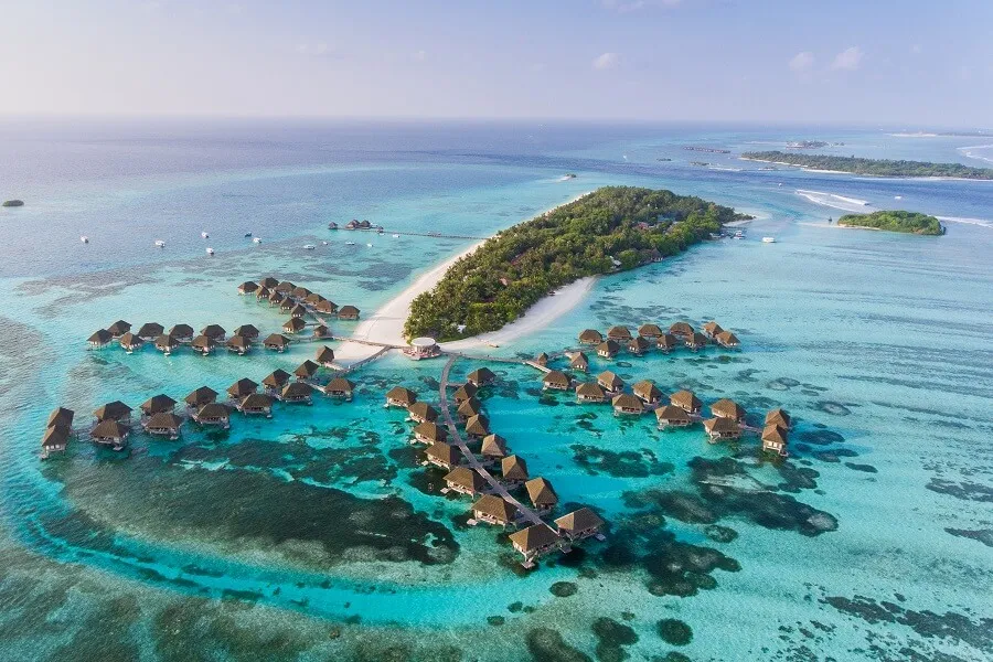 Malediven mit luxuriösen Wasservillen auf türkisblauem Meer, umgeben von grünen Inseln mit weißen Sandstränden. Ein langer Steg verbindet die Villen mit einer Hauptinsel, Boote sind im Wasser verstreut.