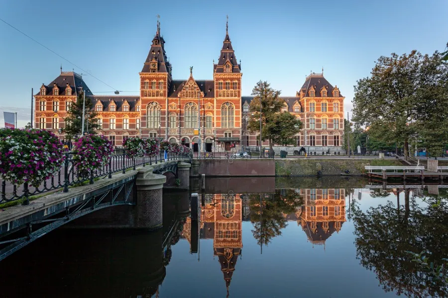 Museumplein in Amsterdam Zuid, Niederlande, im Frühling. Ein zentraler Platz mit Rasenflächen, Bäumen und einem großen 'I Amsterdam'-Schriftzug im Vordergrund. Dahinter sind das Rijksmuseum und das Concertgebouw zu sehen, umgeben von einem lebhaften Stadtbild.