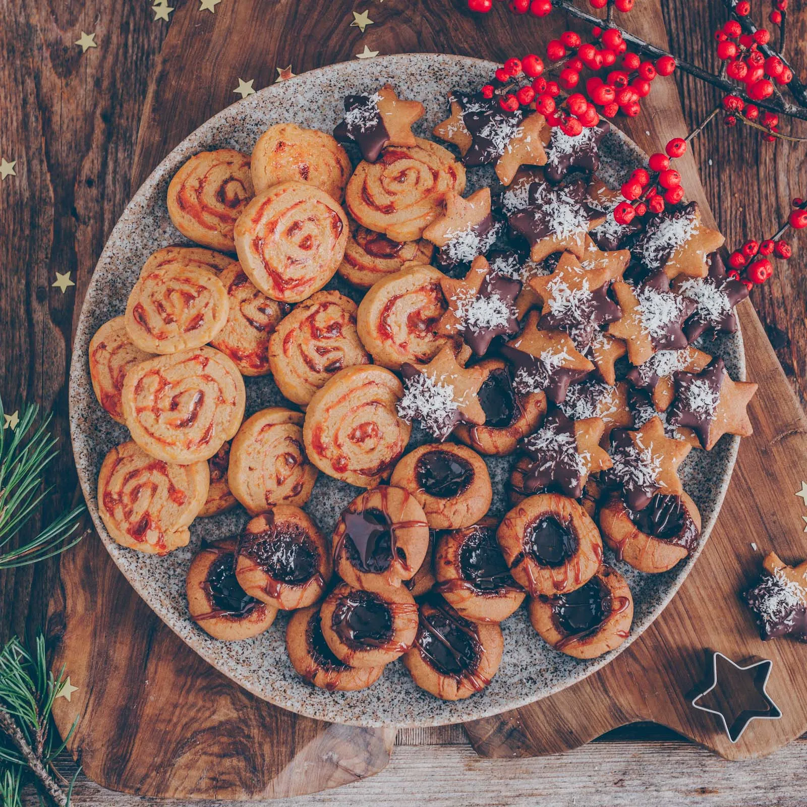 Nahaufnahme einer festlichen Auswahl an veganen Plätzchen, darunter mit Puderzucker bestäubte Kokos-Sterne und gefüllte Thumbprint Cookies, bereit zum Servieren
