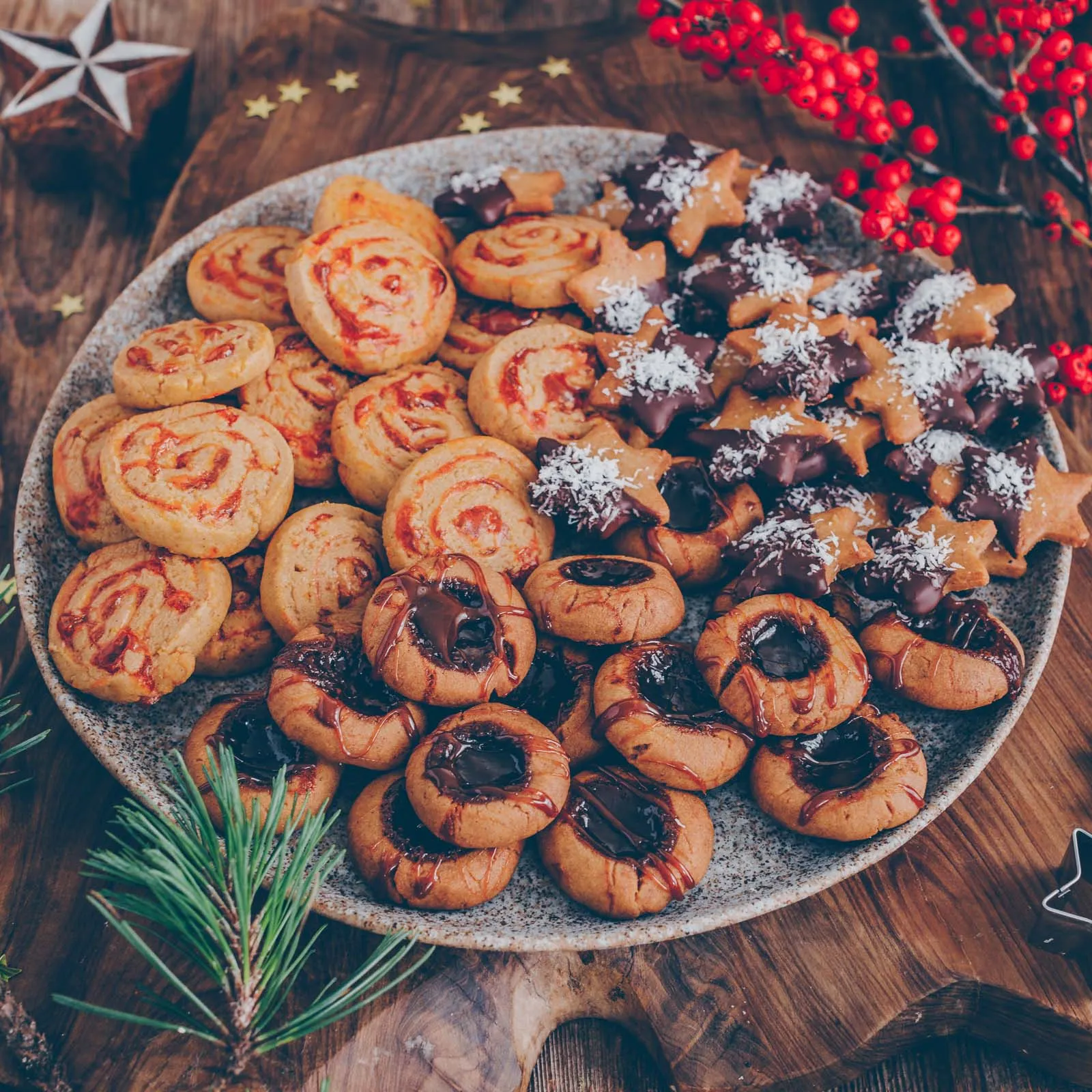 Nahaufnahme einer Mischung aus frisch gebackenen veganen Plätzchen auf einem Holzbrett, darunter Kokos-Sterne, Karamell-Thumbprints und Marmeladen-Rollen