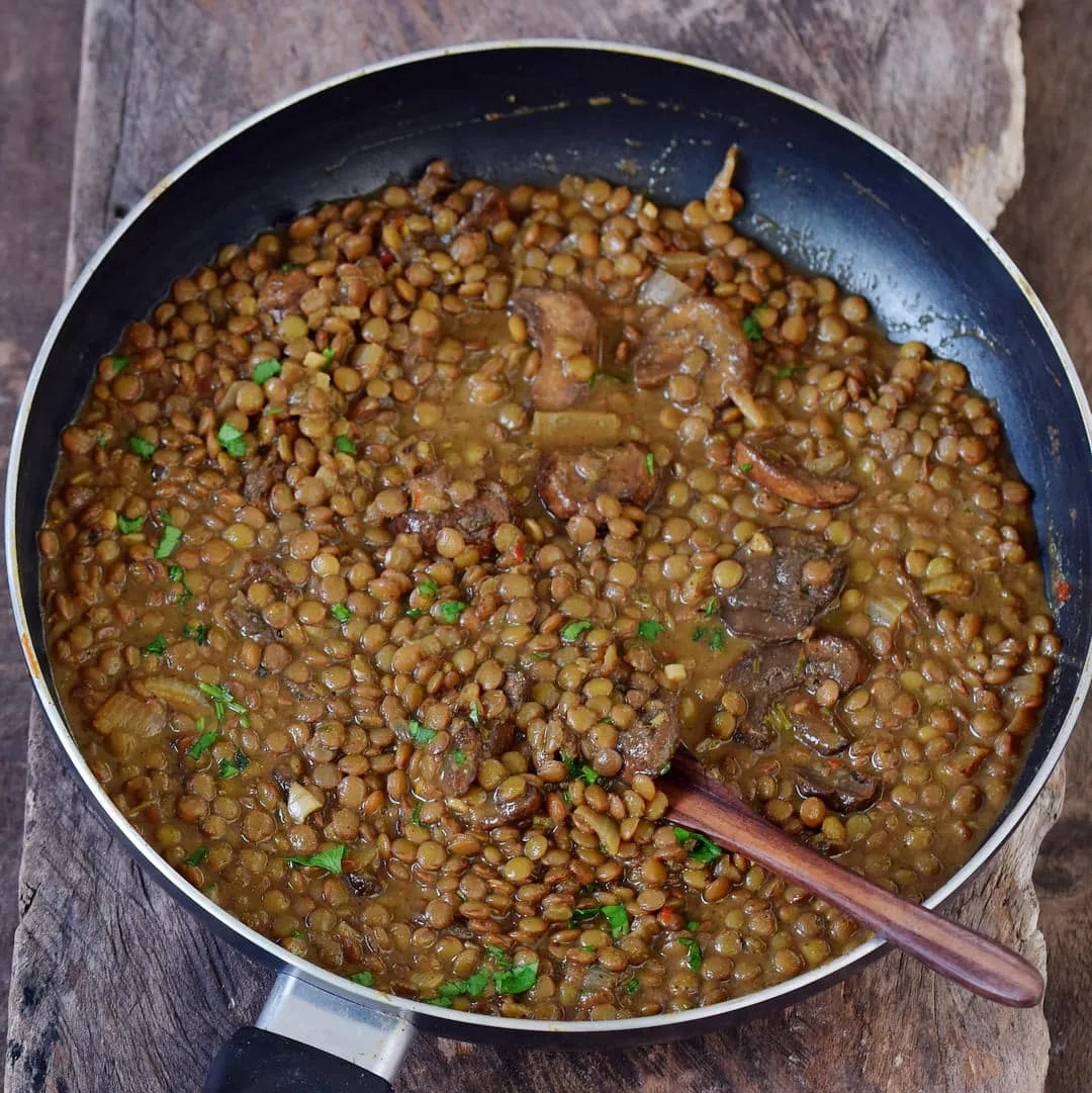 Nahaufnahme einer veganen Linsenpfanne mit Pilzen und Kräutern, angerichtet in einer rustikalen Pfanne, bereit zum Servieren.