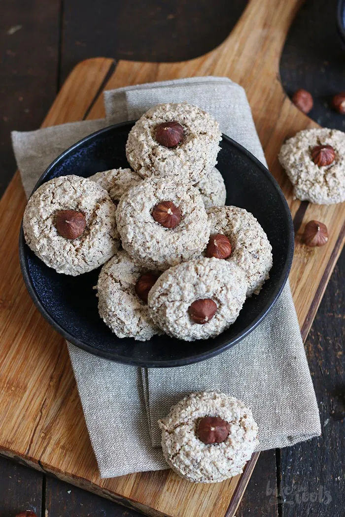 Nahaufnahme von frisch gebackenen Haselnussmakronen auf einem Backblech, die Oberfläche ist leicht gerissen und goldbraun. Im Hintergrund sind weitere Makronen und Backutensilien unscharf zu erkennen.