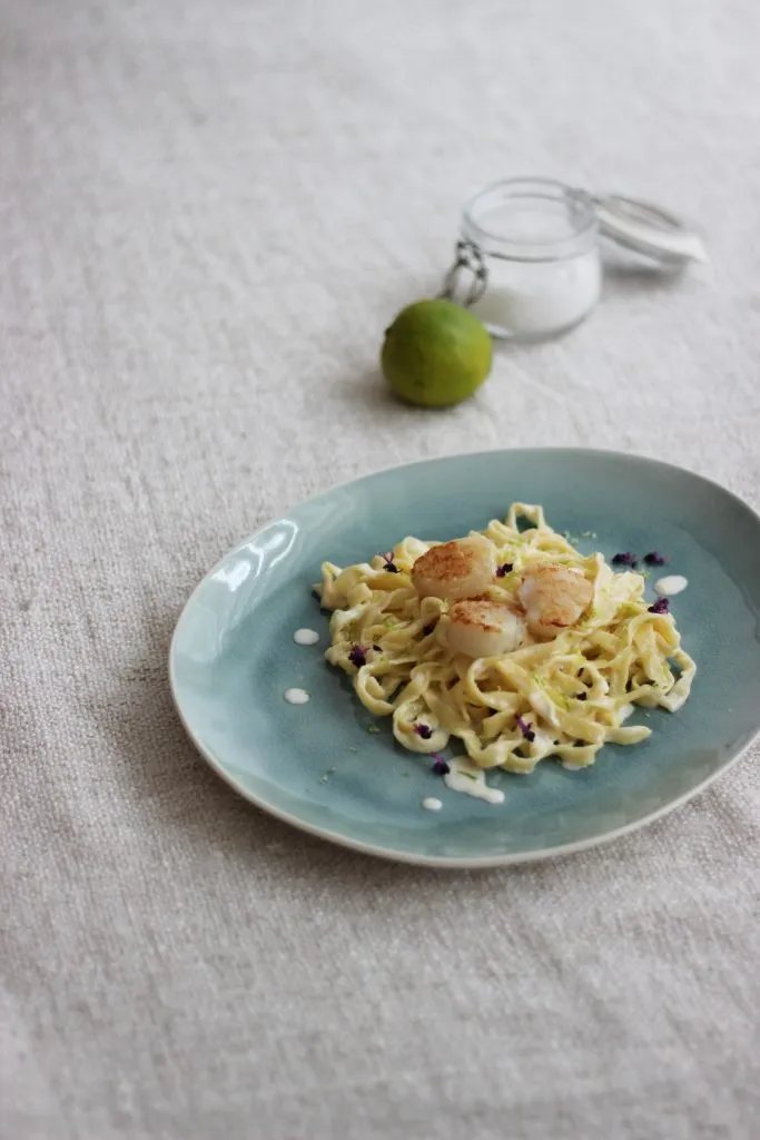 Nahaufnahme von hausgemachten Tagliatelle mit glänzender Limettensauce und perfekt gebratenen Jakobsmuscheln, angerichtet auf einem Teller.