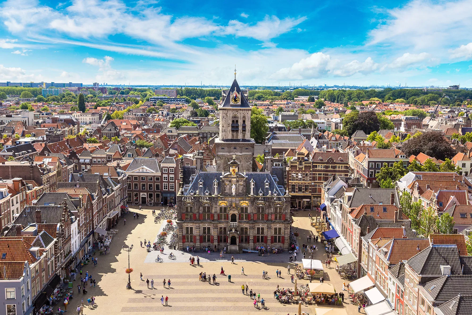 Panoramablick auf Delft mit Fokus auf das Stadhuis im Sommer, umgeben von blühenden Blumen und lebhaften Besuchern.