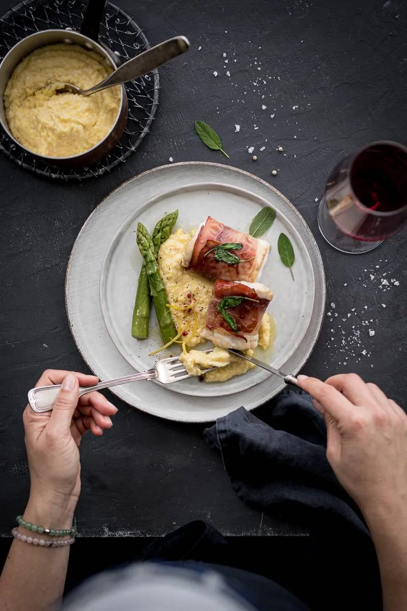 Professionelles Foto von Zander Saltimbocca, das die feine Balance zwischen dem Fisch, dem Speck und der Polenta hervorhebt, serviert mit saisonalem Gemüse.