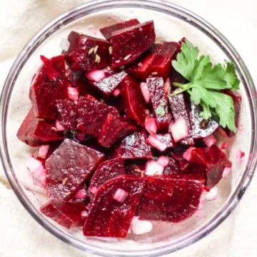 Rote Beete Salat mit gekochten, in Scheiben geschnittenen roten Rüben, fein gewürfelten Zwiebeln und einem grünen Petersilienblatt als Garnitur in einer Glasschüssel.
